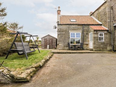 An outdoor area with a stone cottage a wooden swing on grass a stone shed and a table with two chairs at Hawthorn Dale Cottage Whitby