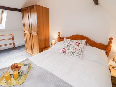 A bedroom with a wooden bed with white bedding and a floral pillow two wooden wardrobes two bedside tables with lamps and a tray with croissants and juice on the bed at Hawthorn Dale Cottage in Whitby