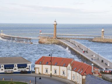 A harbor with two stone piers and lighthouses extending into the sea with buildings and a road in the foreground at Hawthorn Dale Cottage in Whitby