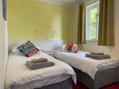A bedroom with two single beds side by side each with a colorful cushion and folded towels a window with beige curtains a bedside table and wall lights at The Wheel @ Canllefaes Penparc near Cardigan