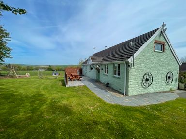 A green stone house with a slate roof and black wagon wheels on the side a picnic table on a stone patio and a grassy lawn with a playground at The Wheel @ Canllefaes in Penparc near Cardigan