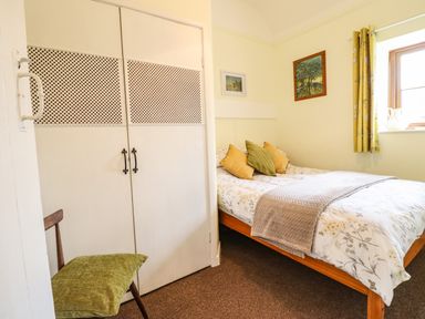 A bedroom with a bed, pillows, a blanket, a chair with a cushion, wardrobe, two paintings, and a window with curtains at Coed Y Glyn in Llanuwchllyn near Bala
