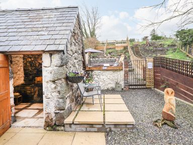 An outdoor patio area with stone building a wooden door metal chairs and a carved wooden owl at Coed Y Glyn in Llanuwchllyn near Bala