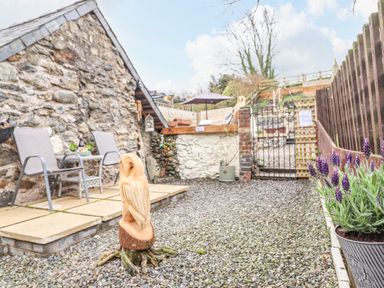 An outdoor stone patio area with two chairs and a table next to a stone building and a wooden owl carving on gravel at Coed Y Glyn in Llanuwchllyn near Bala