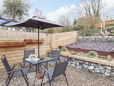 An outdoor patio area with a glass table, four chairs, a black umbrella, and wooden planters on a gravel ground at Coed Y Glyn in Llanuwchllyn near Bala