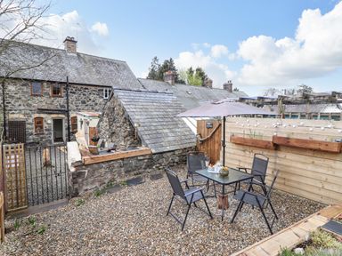 An outdoor patio area with a glass table and four chairs under an umbrella next to stone buildings at Coed Y Glyn in Llanuwchllyn near Bala