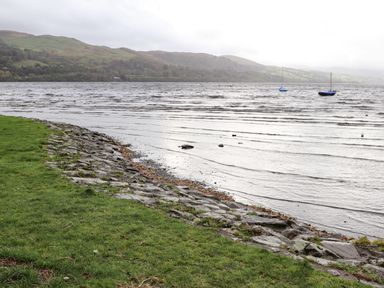 A lakeside view with grassy shore and rocks with boats on the water at Coed Y Glyn in Llanuwchllyn near Bala
