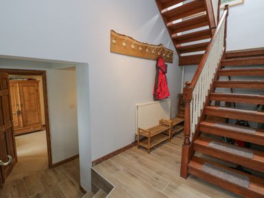 A hallway with wooden stairs a coat rack with a red jacket a wooden shoe rack and an open door showing a room with a wooden wardrobe at Pennant Tigi Uchaf in Dinas Mawddwy