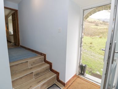 A hallway with stairs leading up and an open door revealing a grassy outdoor area with boots outside at Pennant Tigi Uchaf in Dinas Mawddwy