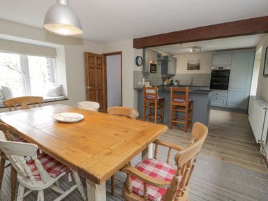 A dining area with a wooden table and six chairs next to a kitchen with two wooden stools at Pennant Tigi Uchaf in Dinas Mawddwy