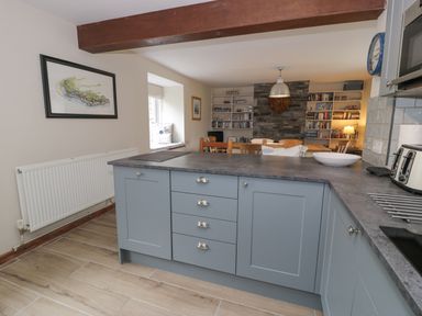 A kitchen area with gray cabinets and countertop overlooking a dining room with wooden chairs and bookshelf at Pennant Tigi Uchaf in Dinas Mawddwy