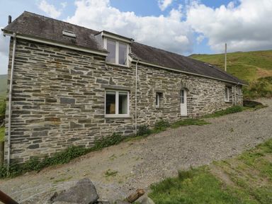 Stone house with multiple windows and a white door on a gravel path with green hills and cloudy sky at Pennant Tigi Uchaf in Dinas Mawddwy