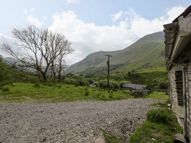 A gravel path beside a stone building with green hills, a leafless tree, and a wooden utility pole at Pennant Tigi Uchaf in Dinas Mawddwy