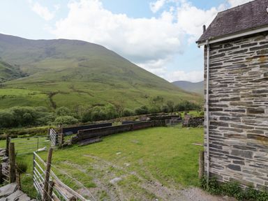 A stone building with a fenced yard and green hills in the background at Pennant Tigi Uchaf in Dinas Mawddwy