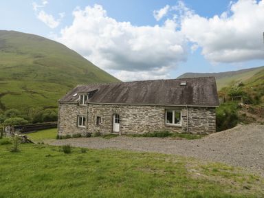 A stone house with a dark roof surrounded by green hills under a partly cloudy sky at Pennant Tigi Uchaf in Dinas Mawddwy