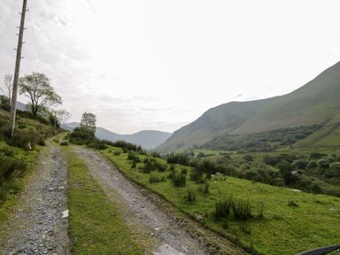 A gravel path with grass on the sides leading through a green hilly landscape with trees and a utility pole at Pennant Tigi Uchaf in Dinas Mawddwy