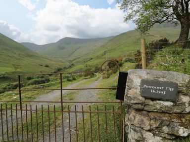 A metal gate with a stone post and a sign reading Pennant Tigi Uchaf with a gravel path and green hills in the background at Pennant Tigi Uchaf in Dinas Mawddwy