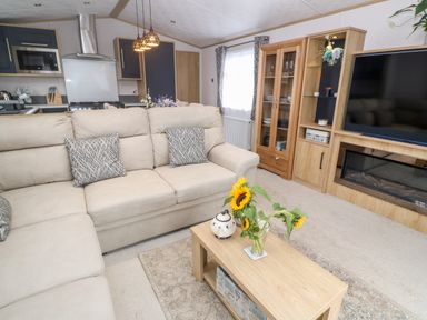 A living room with a beige sofa coffee table with sunflowers and a vase wooden cabinet television and kitchen in the background at Chantry Lodge Hendra Croft near Goonhavern