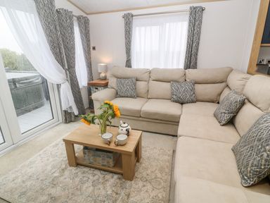 A living room with a beige sectional sofa a wooden coffee table with sunflowers and teapot set near windows with curtains at Chantry Lodge Hendra Croft near Goonhavern
