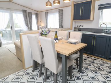 A dining area with a wooden table, four upholstered chairs, a kitchen with blue cabinets, and a living room with curtains at Chantry Lodge Hendra Croft near Goonhavern