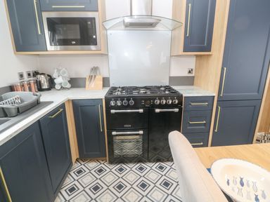 A kitchen with a black stove, blue cabinets, microwave, kettle, knife set, and drying rack with cups at Chantry Lodge in Hendra Croft near Goonhavern