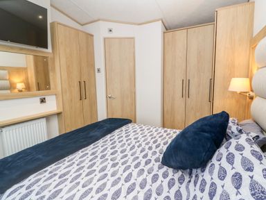 A bedroom with a bed covered in patterned bedding and blue pillows two wooden wardrobes a door a wall-mounted TV and a bedside lamp at Chantry Lodge in Hendra Croft near Goonhavern