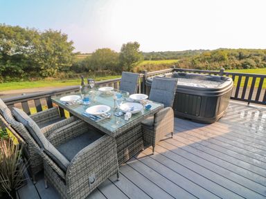 An outdoor dining table with four chairs and place settings beside a hot tub on a deck with a view of trees and fields at Chantry Lodge in Hendra Croft near Goonhavern