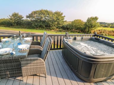 An outdoor deck with a dining table set with plates glasses and a bottle next to a hot tub overlooking greenery at Chantry Lodge in Hendra Croft near Goonhavern