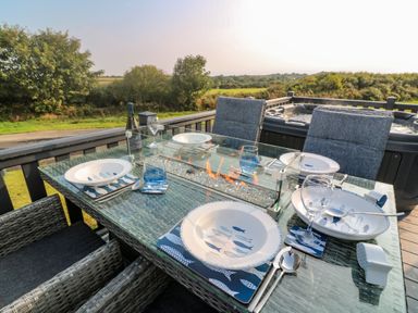 An outdoor glass dining table set with plates bowls glasses utensils and a central fire feature with cushioned chairs and a hot tub on a wooden deck at Chantry Lodge in Hendra Croft near Goonhavern
