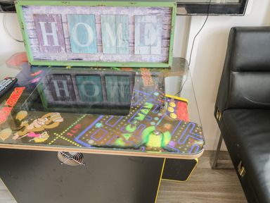 A vintage arcade game table with pac-man and donkey kong graphics with a framed home sign on top and a black leather chair next to it at Wyvill House in Cayton
