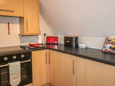 A kitchen corner with wooden cabinets black countertop a red toaster a black bread box a cookbook and an oven with a green oven mitt at Hillside in Gunnislake