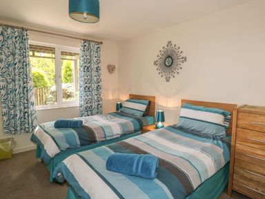 A bedroom with two single beds with striped bedding a wooden chest of drawers and a window with patterned curtains at Hillside in Gunnislake