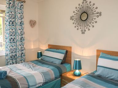 A bedroom with two single beds striped bedding bedside table with lamp and window with patterned curtains at Hillside in Gunnislake