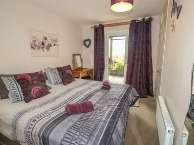 A bedroom with a large bed with patterned bedding and pillows two rolled towels on the bed purple curtains on a glass door wooden nightstand with a mirror and heart decoration on the wall at Hillside in Gunnislake