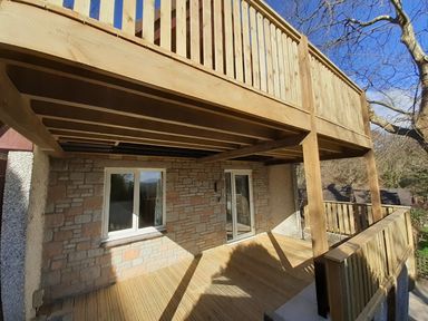 A wooden deck with railings and support beams outside a stone house wall with a window and a glass door at Hillside in Gunnislake