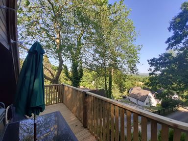 A wooden balcony with a glass table and closed umbrella overlooking trees and houses at Hillside in Gunnislake