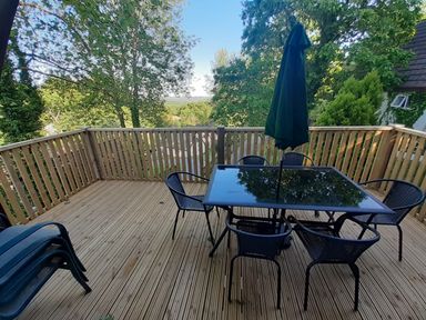 An outdoor wooden deck with a glass table and chairs and a closed umbrella surrounded by trees at Hillside in Gunnislake