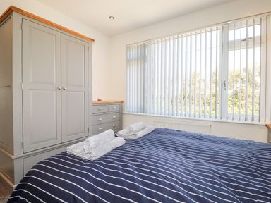 A bedroom with a bed with striped bedding towels on the bed a large window with vertical blinds and a gray wardrobe with drawers at Seawynds in Crantock