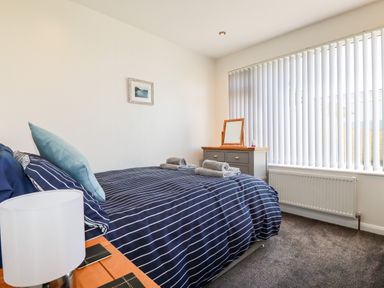 A bedroom with a bed striped blue bedding and pillows beside a wooden nightstand with a lamp next to a window with vertical blinds and a dresser with a mirror at Seawynds in Crantock