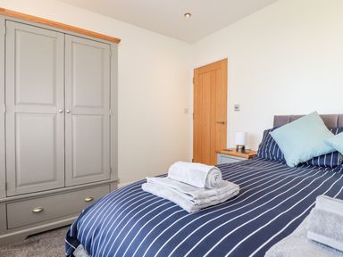 A bedroom with a bed with striped bedding folded towels headboard bedside table with lamp wooden door and grey wardrobe at Seawynds in Crantock