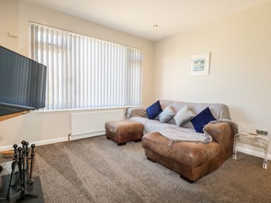 A living room with a brown leather sofa and ottoman a wall-mounted television and vertical blinds on the window at Seawynds in Crantock