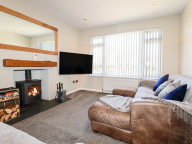A living room with a wood stove fireplace a TV on the wall a leather sofa with cushions and a large window with vertical blinds at Seawynds in Crantock