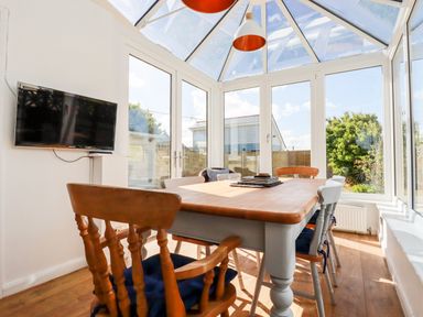 A dining area with wooden chairs and table under a glass roof with a wall mounted TV at Seawynds in Crantock