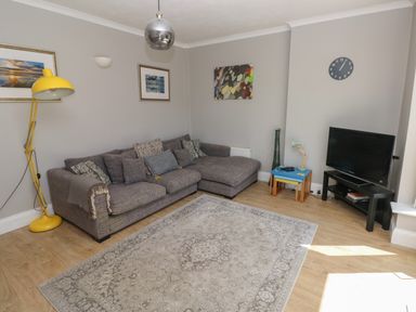 A living room with a gray sofa yellow floor lamp a blue side table and a television on a black stand at Pinewood in Saundersfoot
