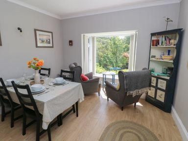 A dining area with a table set for six and a seating area with two armchairs next to a window at Pinewood in Saundersfoot