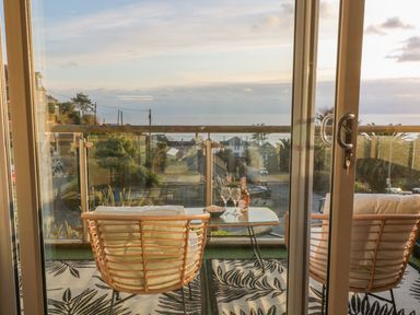 A balcony with two cushioned chairs a glass table with a bottle and glasses overlooking houses and the sea at A Stone's Throw in Downderry