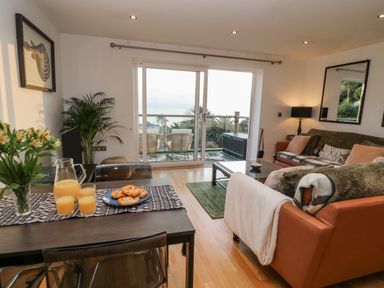 A living room with a dining table set with juice and pastries and a seating area with sofas and a coffee table at A Stone's Throw in Downderry