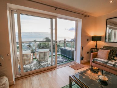A living room with a glass coffee table a brown couch and sliding doors leading to a balcony at A Stone's Throw in Downderry
