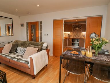 A living room with a leather sofa and a dining table with flowers and juice near a kitchen with wooden doors at A Stone's Throw in Downderry