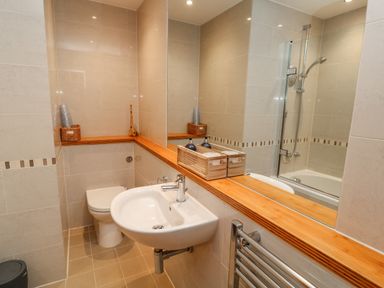 A bathroom with a white sink, toilet, bathtub with shower, and wooden shelf at A Stone's Throw in Downderry